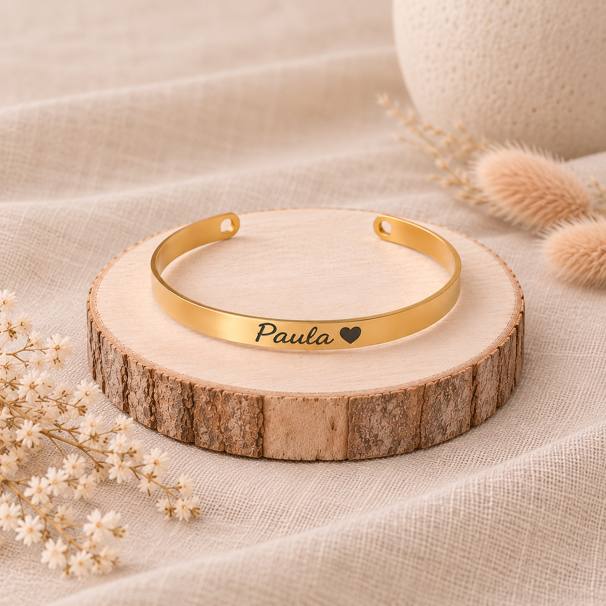 Brazalete rígido Imara dorado personalizado con nombre Paula y corazón grabado, sobre rodaja de madera con flores secas